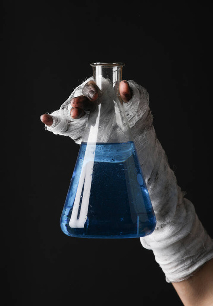 Mama 's hand gewikkeld in een verband houdt een laboratoriumfles vast met een elixer geïsoleerd op een zwarte achtergrond. Halloween concept - Foto, afbeelding
