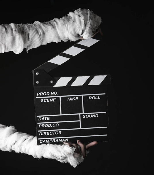 Mummy's hand wrapped in bandage holds movie clapperboard isolated on black background. Halloween, movie concept - Foto, imagen