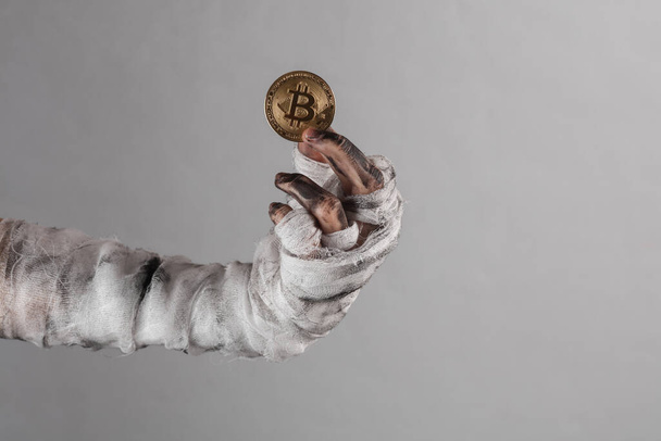 Mummy's hand wrapped in bandage holds bitcoin isolated on gray background. Halloween, business concept - Foto, imagen
