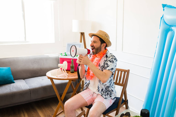 Aantrekkelijke gelukkige man met behulp van een megafoon schreeuwen in de woonkamer terwijl u wilt reizen naar het strand op vakantie  - Foto, afbeelding