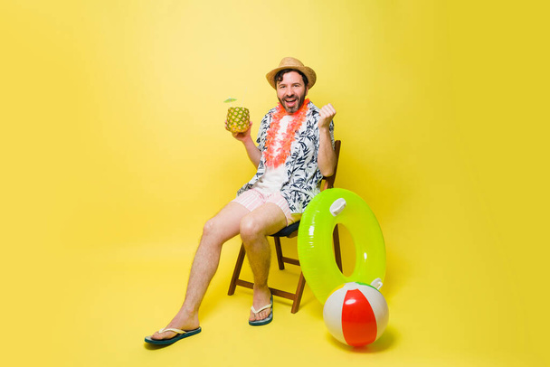 Attractive happy man wearing shorts and a hat during his summer vacations enjoying a pineapple drink against a yellow background - Photo, Image