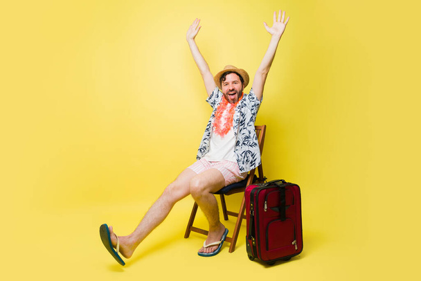 Portrait of an excited man having fun celebrating going on a summer travel to the beach with a suitcase in front of a yellow background - Foto, Imagem
