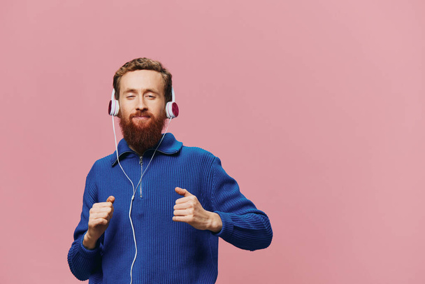 Portrait d'un homme roux portant des écouteurs souriant et dansant, écoutant de la musique sur fond rose. Un hipster avec une barbe. Photo de haute qualité - Photo, image