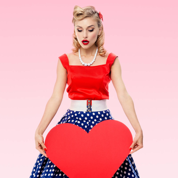 Woman holding heart symbol, dressed in pin-up style dress with polka dot, over pink background. Caucasian blond model posing in retro fashion and vintage concept studio shoot. - Photo, image