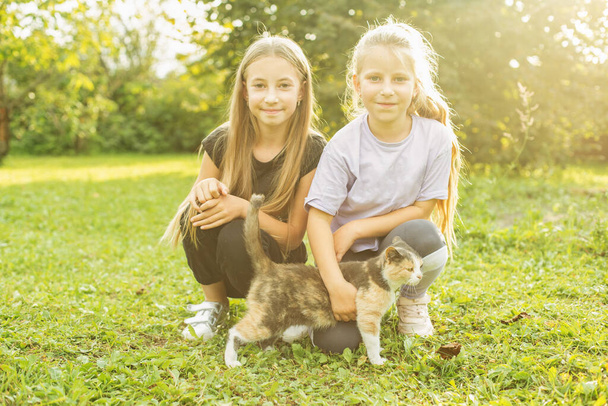 Kaksi lasta leikkii kotimaisen kissan kanssa talon pihalla. Aurinkoinen päivä. Lapsuuden ja lemmikkieläinten käsite. - Valokuva, kuva