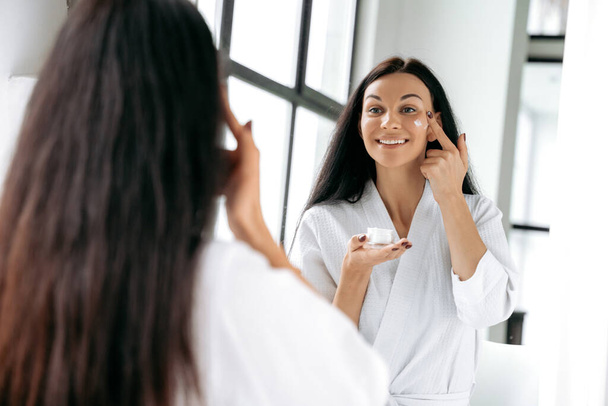 Nutrition and hydration of the skin. Gorgeous caucasian brunette long haired woman in a white bathrobe stands in the bathroom in front of a mirror, applying moisturizer to face, smiling - Fotoğraf, Görsel