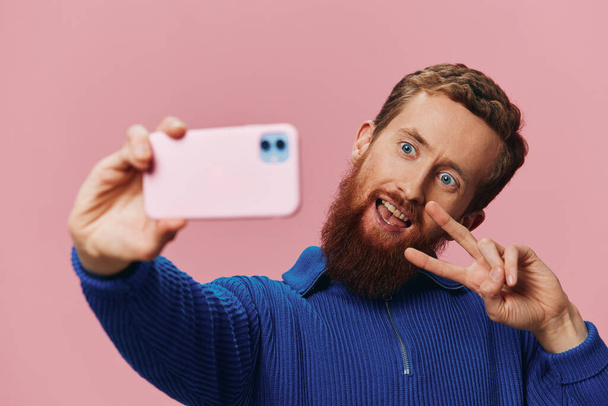 Портрет рыжеволосого человека с телефоном в руке, делающего селфи и фотографии на телефоне с улыбкой на розовом фоне, блоггер. Высокое качество фото - Фото, изображение