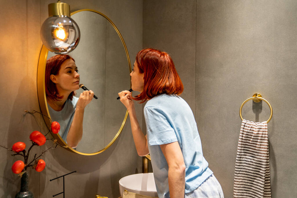 Chica pelirroja mirando en el espejo aplicando maquillaje todos los días en la cara con cepillo. Mujer joven de pie en el baño usando polvo compacto para cubrir defectos. Adaptación a los estándares de belleza de la sociedad - Foto, Imagen