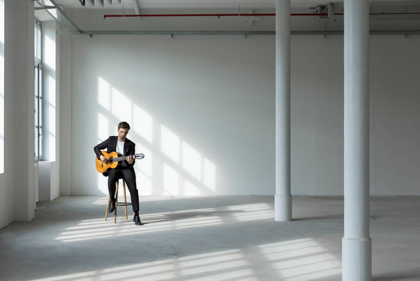Portrait of a young boy playing the guitar. He is wearing a black suit. Interior of a renovated former factory. - Foto, Bild