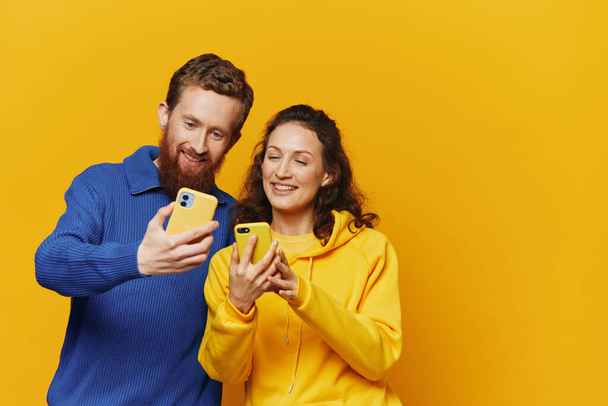 Hombre y mujer pareja sonriendo alegremente con el teléfono en la mano redes sociales ver fotos y videos, sobre fondo amarillo, símbolos signos y gestos de la mano, freelancers familiares. Foto de alta calidad - Foto, Imagen
