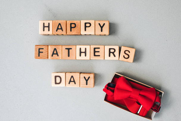 Szczęśliwego dnia ojców. Kontekst, karta gratulacyjna. Drewniane kostki z czarnymi literami układane są na szarym betonowym tle z czerwonymi akcesoriami męskimi, muszką i szelkami. Układy płaskie - Zdjęcie, obraz