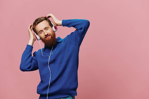 Portrait of a redheaded man wearing headphones smiling and dancing, listening to music on a pink background. A hipster with a beard. High quality photo - Photo, Image