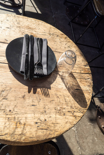 A plate with a rag napkin and cutlery. Two empty upturned glasses on an old wooden textured table cast a shadow from the midday sun - Foto, Imagem