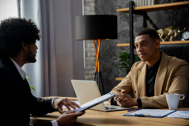 アフリカ系アメリカ人のビジネスマンがテーブルに座り、仕事を報告する従業員の話を聞く。2人の起業家が新しいスタートアップのために資金を調達する方法を議論している - 写真・画像