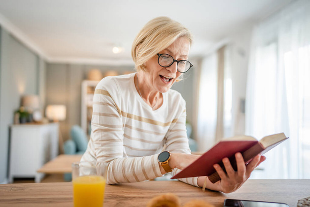 Bir kadın olgun beyaz kadın aydınlık odada modern kitap okuyor gözlük takıyor gerçek bir insan mekanı kopyalıyor. - Fotoğraf, Görsel