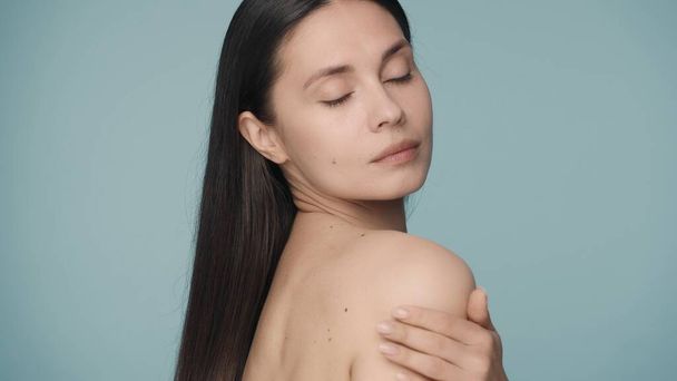 A young woman rubs her shoulder with slow massaging movements with her eyes closed. A seminude brunette woman on a blue background close up. The concept of skin and body care. Beauty and cosmetology - Foto, afbeelding