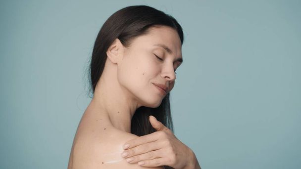 With slow massage movements, the woman applies the cream on her shoulder. The girl with a smile rubs the cream into the skin on a blue background. Close up. The concept of skin and body care. Natural - Foto, imagen