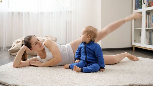 Belle femme souriante allongée sur un tapis avec bébé fils et faisant des exercices de fitness. Santé familiale, mode de vie actif, rôle parental et développement de l'enfant. - Photo, image
