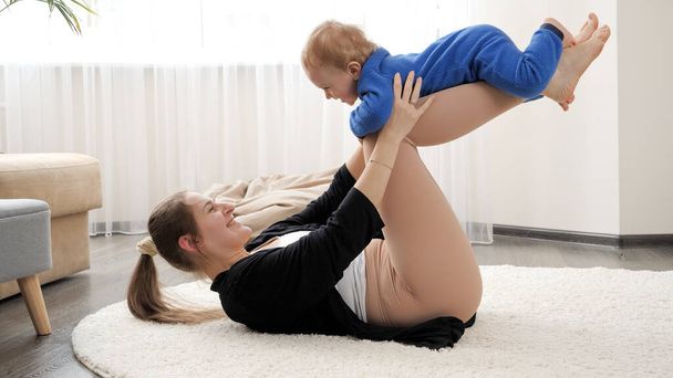 Szczęśliwa uśmiechnięta matka trzymająca i kołysząca synka podczas lekcji fitness i jogi w domu. Rodzinna opieka zdrowotna, aktywny tryb życia, rodzicielstwo i rozwój dziecka. - Zdjęcie, obraz