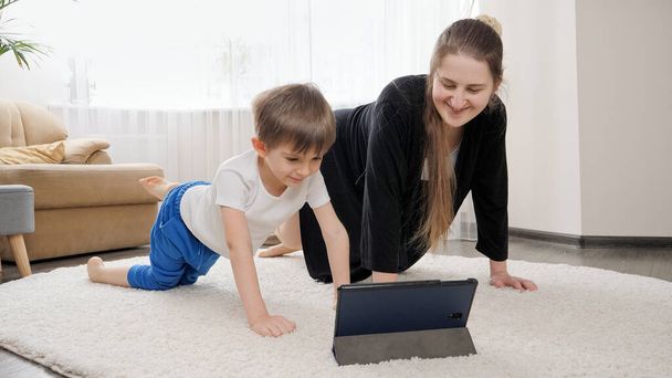 Χαμογελώντας αγόρι με τη μητέρα βλέποντας online μάθημα φυσικής κατάστασης στο tablet και κάνοντας αθλητικές ασκήσεις στο σπίτι. Οικογενειακή υγειονομική περίθαλψη, ενεργός τρόπος ζωής, γονική μέριμνα και ανάπτυξη παιδιών. - Φωτογραφία, εικόνα