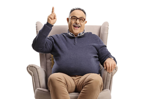 Happy mature man sitting in an armchair and pointing up isolated on white background - Photo, Image