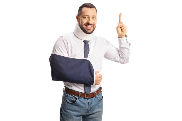 Young injured man with a cervical collar and arm sling pointing up isolated on white background - Fotografie, Obrázek