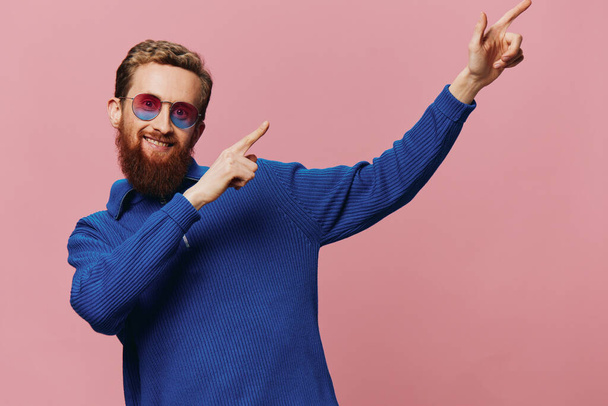 Portret rudego mężczyzny w okularach przeciwsłonecznych, uśmiechniętego i tańczącego, słuchającego muzyki na różowym tle. Hipster z brodą, wskazujący palcem szczęścia. Wysokiej jakości zdjęcie - Zdjęcie, obraz