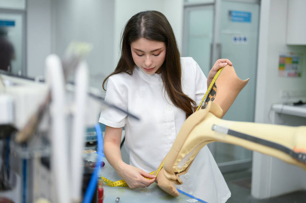 Młoda technik trzymająca protezę nóg i pracująca w laboratorium, Specjalista z zaawansowaną technologią w produkcji protez, Nowa produkcja sztucznych kończyn dla osób niepełnosprawnych - Zdjęcie, obraz