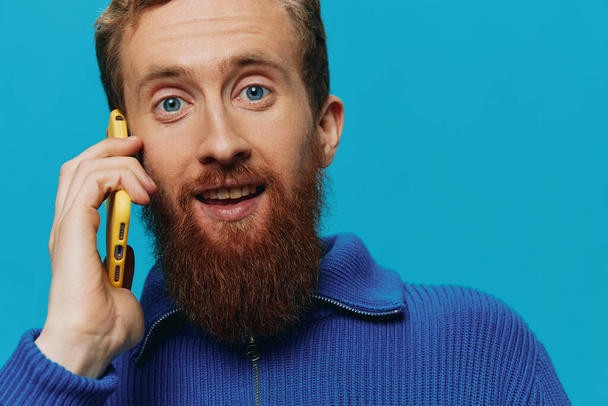 Portret van een man met een telefoon in zijn handen die praat, belt, op een blauwe achtergrond. Communiceren online sociale media, levensstijl. Hoge kwaliteit foto - Foto, afbeelding