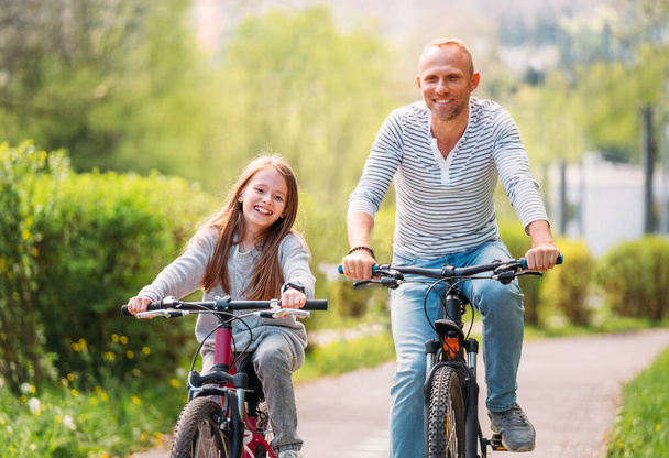 Portraits Smiling father with daughter during summer outdoor bicycle riding. They enjoy togetherness in the summer city park. Happy parenthood and childhood or active sport life concept image. - Photo, Image