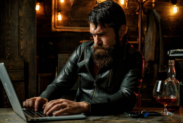 Bearded man drink cognac. Drunk attractive handsome sad pensive man in black jacket, business man drinking whiskey in front of laptop at loft work place - Photo, Image