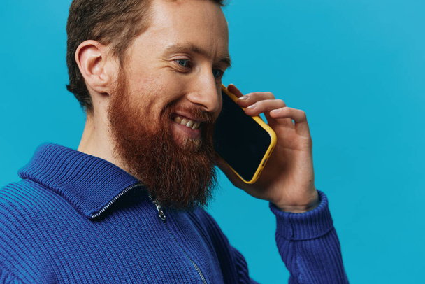 Egy férfi portréja, akinek telefon van a kezében, ránéz és beszél telefonon, kék háttérrel. Kommunikáció online közösségi média, életmód. Kiváló minőségű fénykép - Fotó, kép