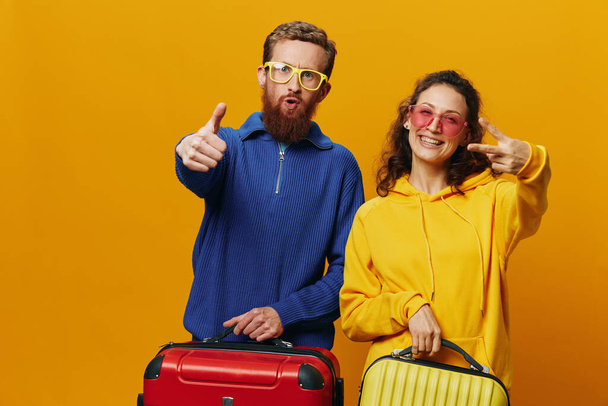 Γυναίκα και άντρας χαμογελαστοί, βαλίτσες στο χέρι με κίτρινη και κόκκινη βαλίτσα χαμογελώντας χαρούμενα και στραβά, κίτρινο φόντο, πηγαίνοντας ταξίδι, οικογενειακό ταξίδι διακοπών, νεόνυμφοι. Υψηλής ποιότητας φωτογραφία - Φωτογραφία, εικόνα