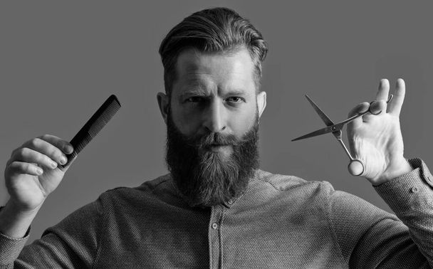 handsome barber man with scissors do hairstyle. man has beard and hairstyle in studio. man hold barber scissors and comb. barber with hairstyle. barber man making hairstyle isolated on grey background - 写真・画像