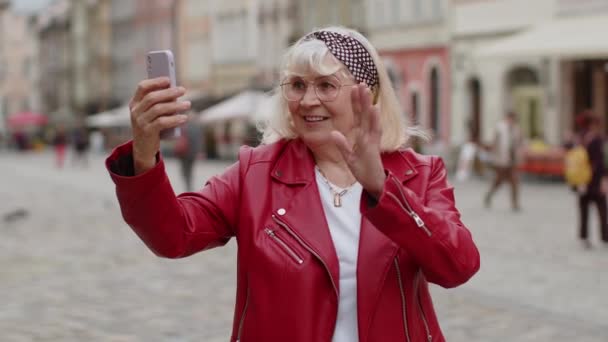Старшая блоггер делает селфи на смартфоне, общается с абонентами через интернет, записывает истории для видеоблога в социальных сетях на открытом воздухе. Пожилая бабушка гуляет по городской улице - Кадры, видео