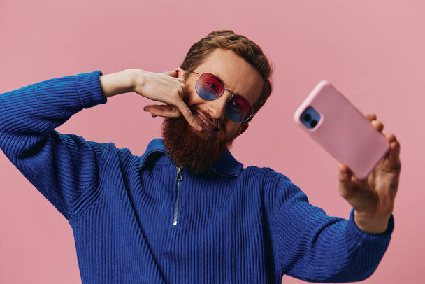 Πορτρέτο ενός κοκκινομάλλη με το τηλέφωνο στο χέρι να βγάζει selfies και φωτογραφίες στο κινητό του με ένα χαμόγελο σε ροζ φόντο, blogger. Υψηλής ποιότητας φωτογραφία - Φωτογραφία, εικόνα