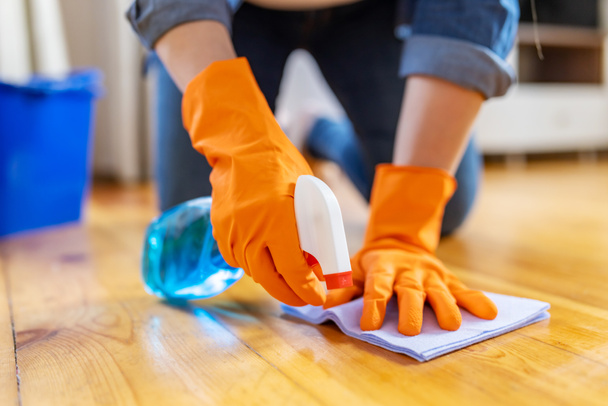 Lastik eldivenli genç bir kadın evin zeminini temizlerken tozunu sprey ve bezle siliyor. - Fotoğraf, Görsel