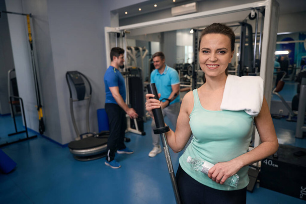 Uśmiechnięta kobieta sportowiec z butelką wody i sztangi stojącej wśród maszyn do ćwiczeń i partnerów fitness na siłowni - Zdjęcie, obraz