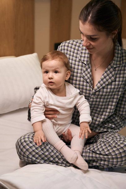 Parent féminin attentionné en pyjama assis avec bébé malade au lit dans la salle - Photo, image