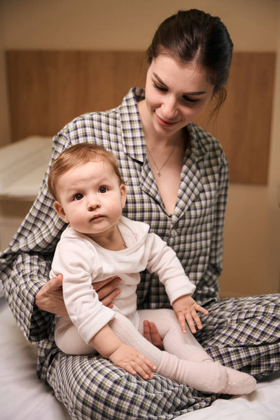 Sonriente joven hembra en pijama sentada con triste bebé hospitalizado en la cama en la sala - Foto, Imagen