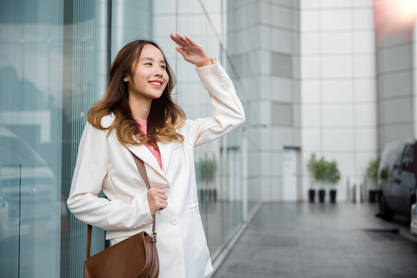 Portrait of confidence young businesswoman standing outside office building in city raise her hand to shade sun UV. Happy woman wearing white suit jacket with brown bag at sunlight outdoors. sun block - Foto, Imagen