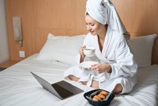 Photo of woman in bathrobe with laptop drinking coffee with croissants on bed in hotel room - Photo, image
