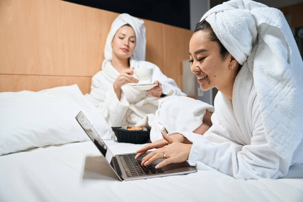 Подружки з ноутбуком і кавою розслабляються в готелі. Дві жінки розмовляють на ліжку в білих халатах
 - Фото, зображення