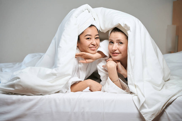 Fénykép két barátnőről, akik a takaró alól mosolyognak. A nők vidáman néznek ki az ágy takarója alól. - Fotó, kép