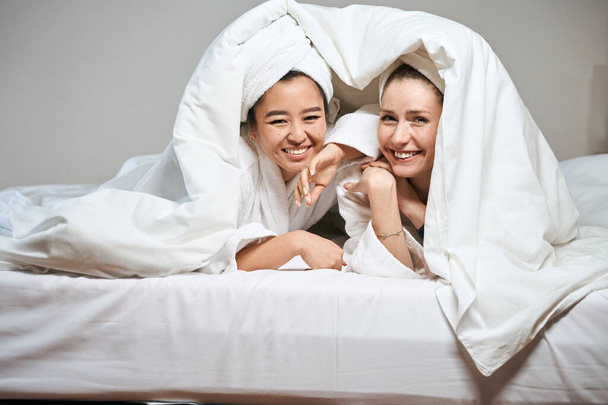 Két vidám barátnő édesen mosolyog a takaró alól. Aranyos hölgyek kukucskálnak ki a takaró alól a szállodai ágyon. - Fotó, kép