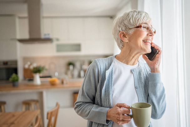 One mature senior senior woman stand at the window at home use mobile phone smartphone to make a call real people daily routine stand in bright room short gray hair wear sweater and eyeglasses - Fotoğraf, Görsel