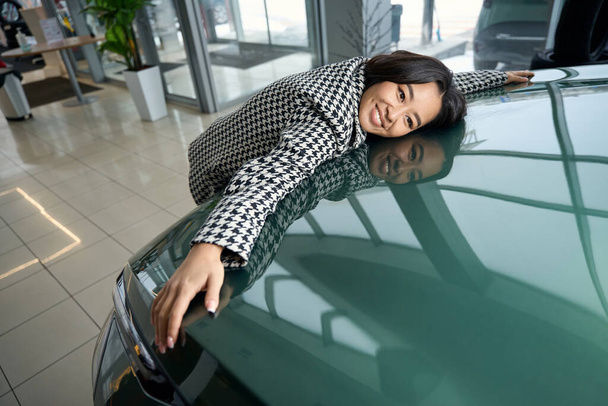 Happy young asian woman hugging the hood of a new car, she is wearing comfortable casual clothes - Photo, Image
