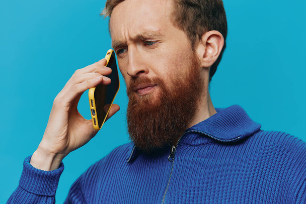 Portret van een man met een telefoon in zijn handen kijkt ernaar en praat erover aan de telefoon, op een blauwe achtergrond. Communiceren online sociale media, levensstijl. Hoge kwaliteit foto - Foto, afbeelding