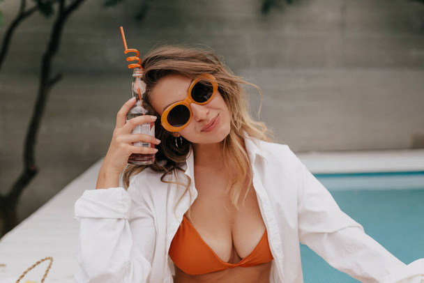 Милая очаровательная леди с волнистыми волосами в оранжевом купальнике и белой рубашке и стильных очках позирует перед камерой и держит летний напиток на фоне бассейна  - Фото, изображение