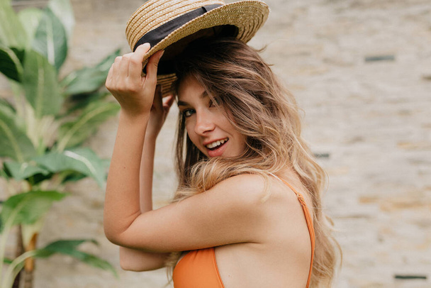 Amazing lovely girl with curly long hair wearing swimsuit waiting for fun in the outdoor pool. Amazing cheerful young woman in hat enjoys vacation in exotic country - 写真・画像